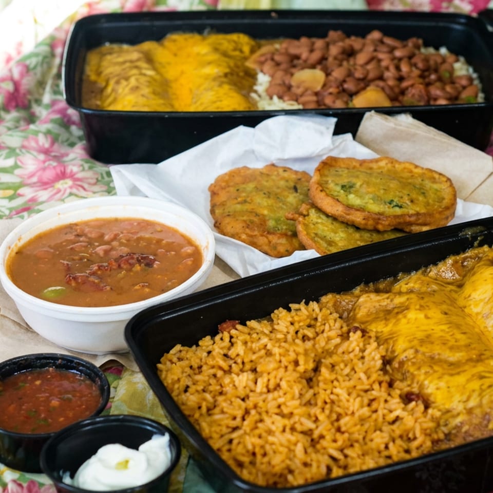 Black takeout trays hold cheese-covered enchiladas, Mexican rice, pinto beans, fried corn cakes, and bowls of beans, salsa, and crema.